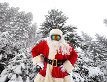 santa wearing ski goggles standing in front of snow covered trees