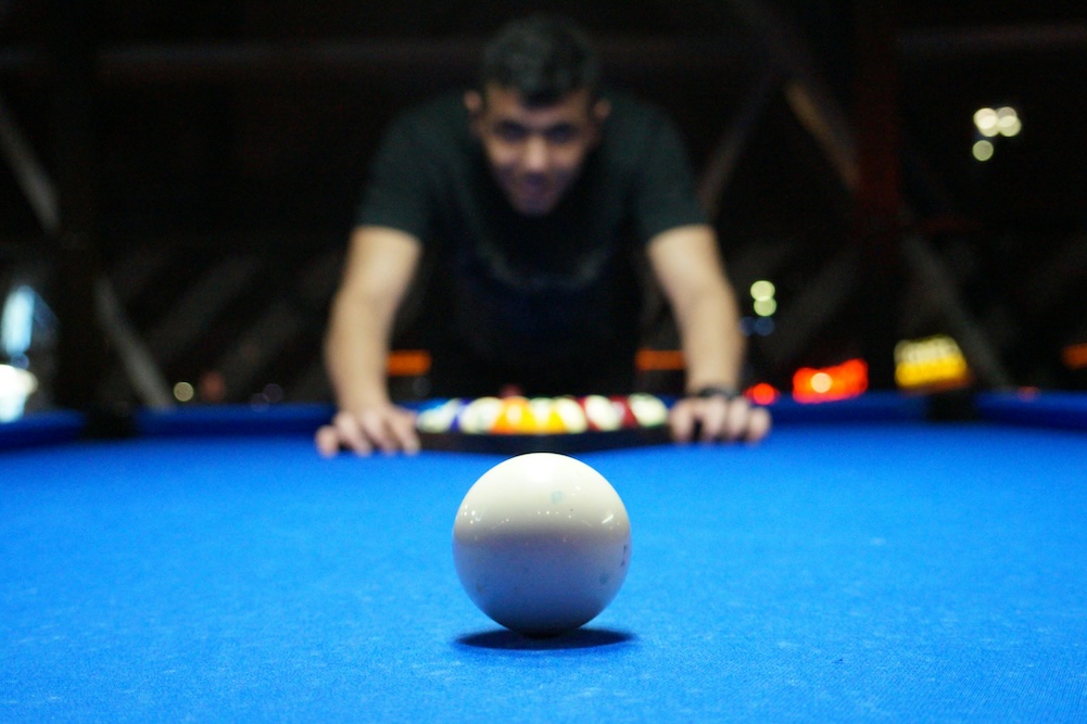 a man in a black t-shirt prepared to break with a cue ball on a blue billiards table at jax food & games